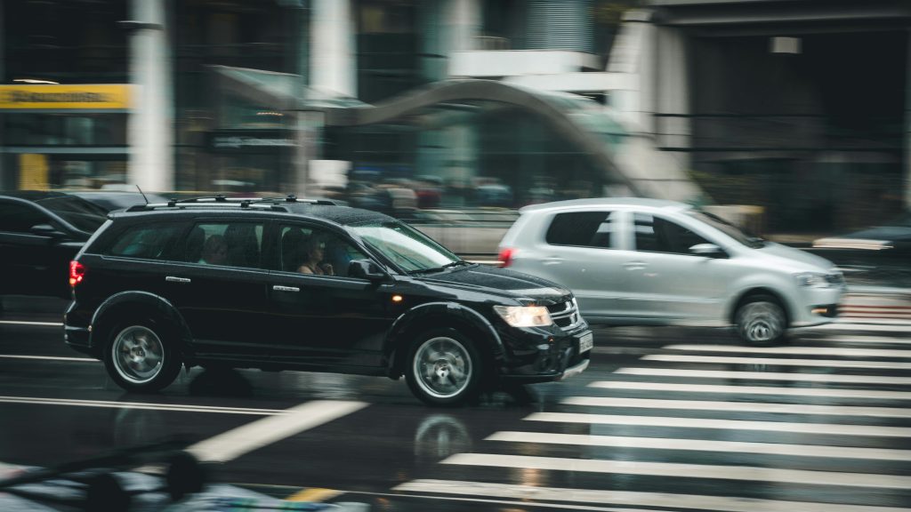Vehicles speeding through a city street with a visible zebra crossing and blurred motion effect.